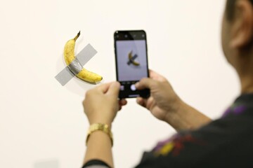 A person is taking a photo of a banana that was priced as a $6 million artwork, in order to establish a personal connection with contemporary art.